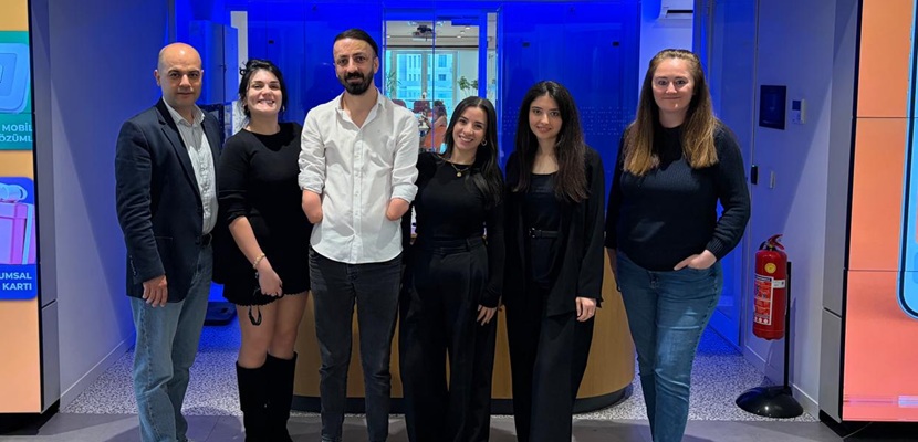 Betimleme: Bülent Kavut ortada bir fuar standı önünde 4 kadın 1 erkek iş arkadaşları ile ayakta yanyana duruken çekilen fotoğraf.